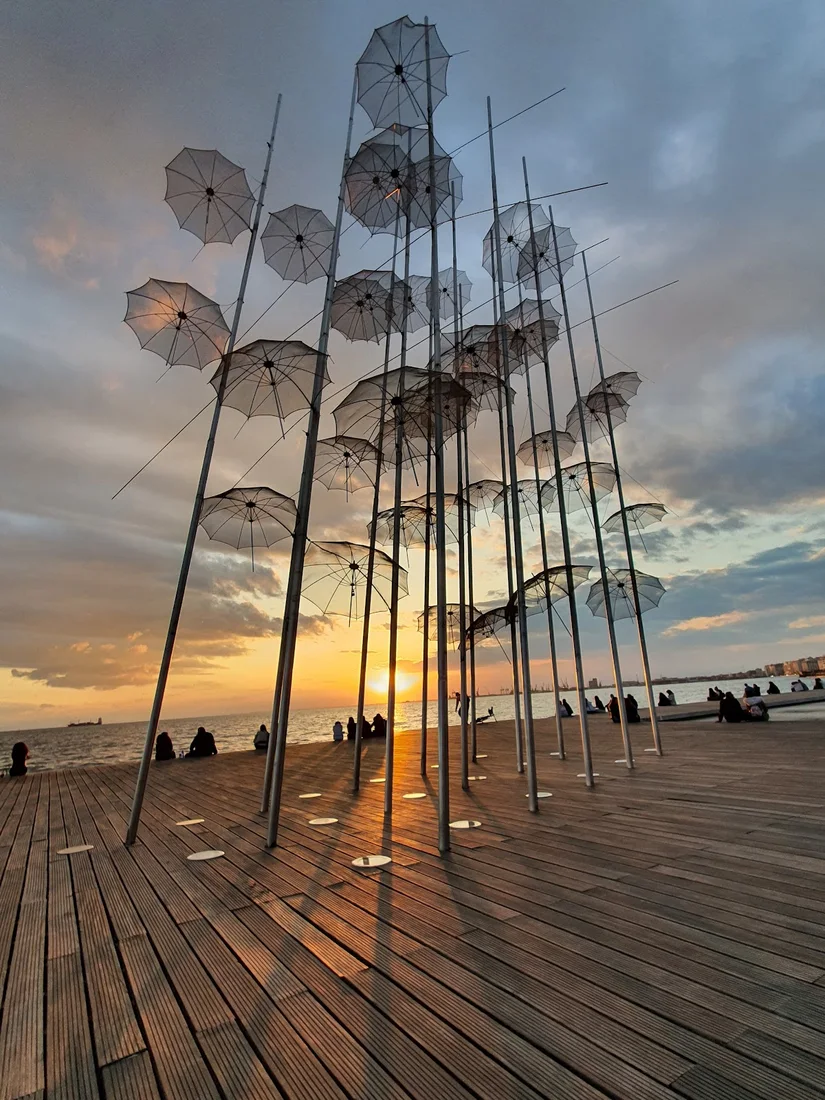 The steel Umbrellas sculpture by Zongolopoulos on the Thessaloniki waterfront, silhouetted against a golden sunset over the gulf