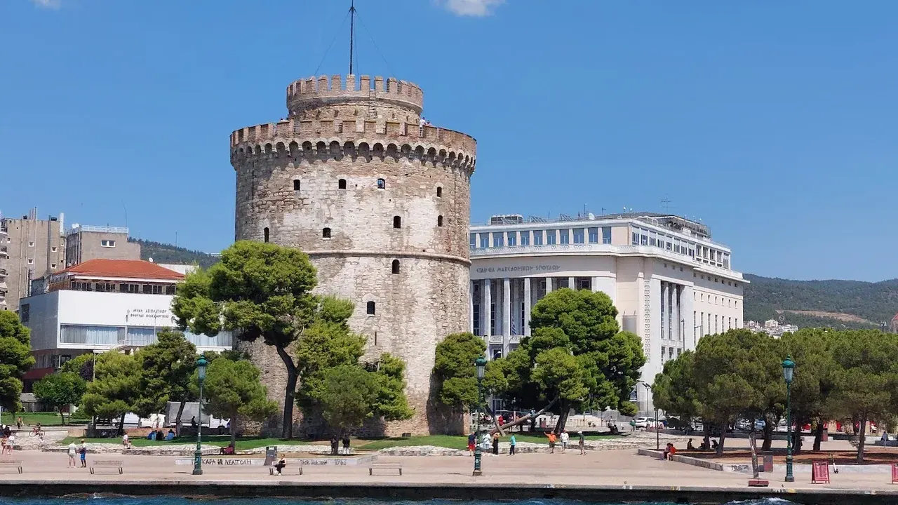 The White Tower of Thessaloniki seen from the water, with its flat paved waterfront promenade and people walking along it