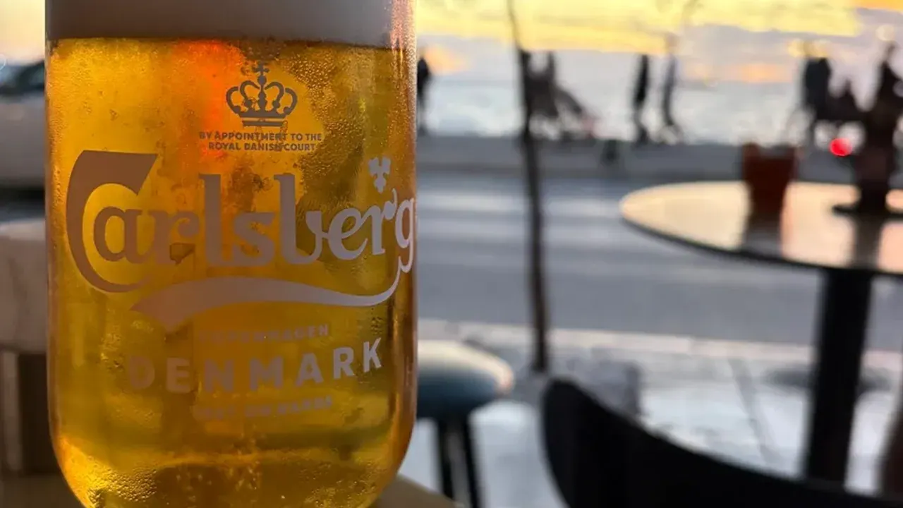A cold beer on ice at a seafront table at sunset, with the promenade and Thermaic Gulf in the background near Thessaloniki