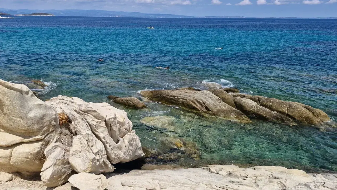 Weathered granite boulders and clear turquoise water with swimmers offshore on a Halkidiki beach
