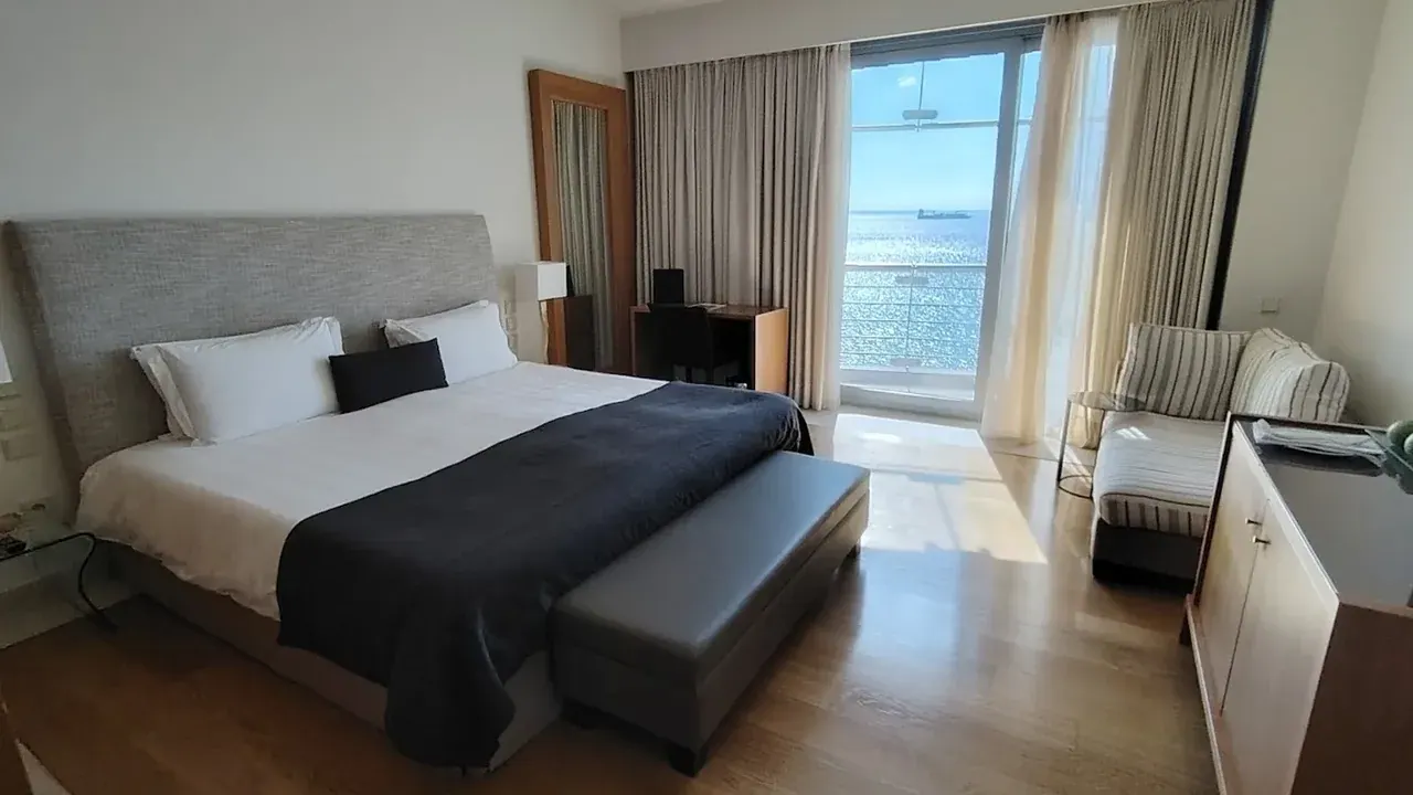 A five-star hotel suite with floor-to-ceiling windows opening onto a balcony over the Thermaic Gulf, a cargo ship visible on the horizon, Thessaloniki