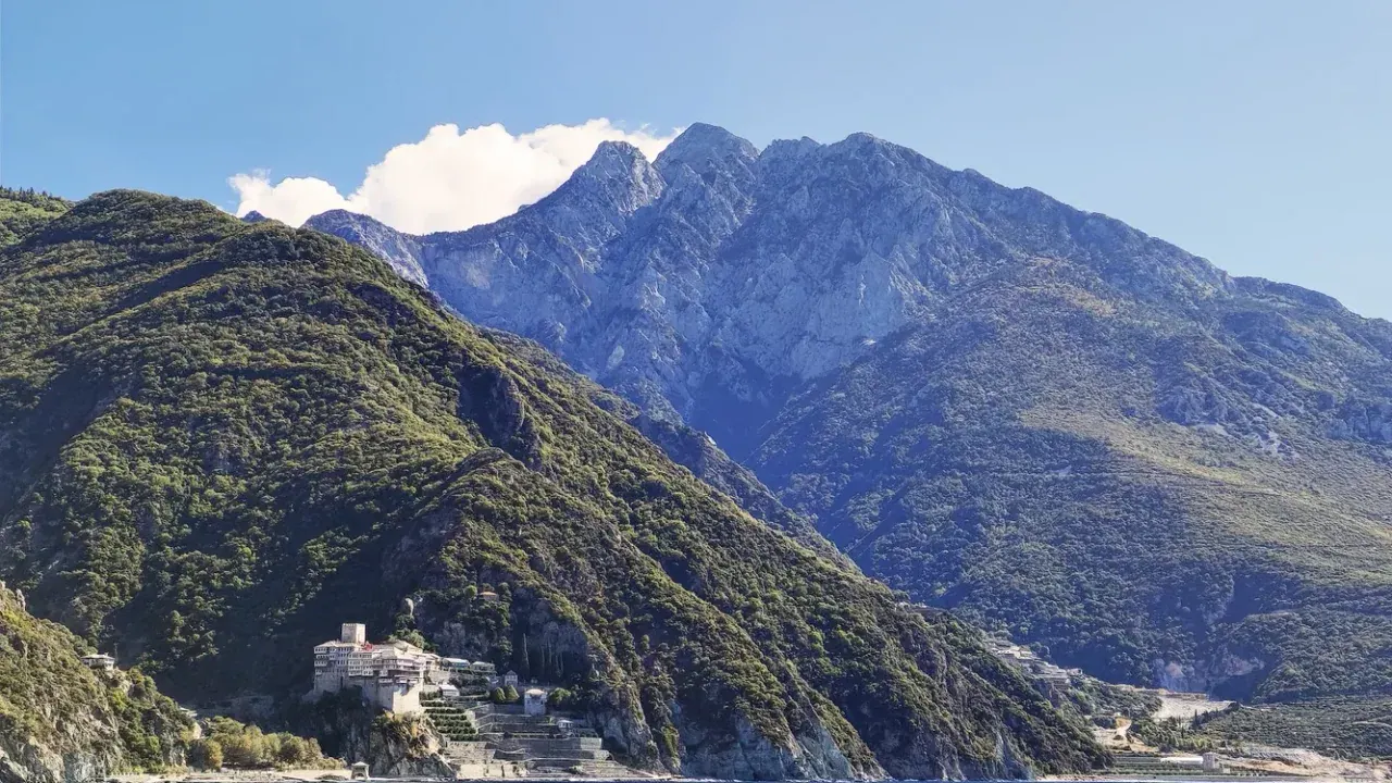 The peak of Mount Athos rising above the forested peninsula with a monastery at the shoreline, seen from the sea