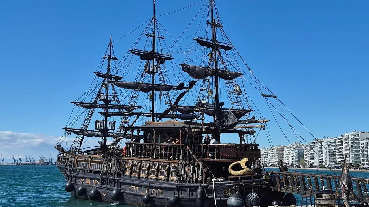 A tourist pirate-ship boat moored on the Nea Paralia waterfront in Thessaloniki, with the flat promenade and city skyline behind it