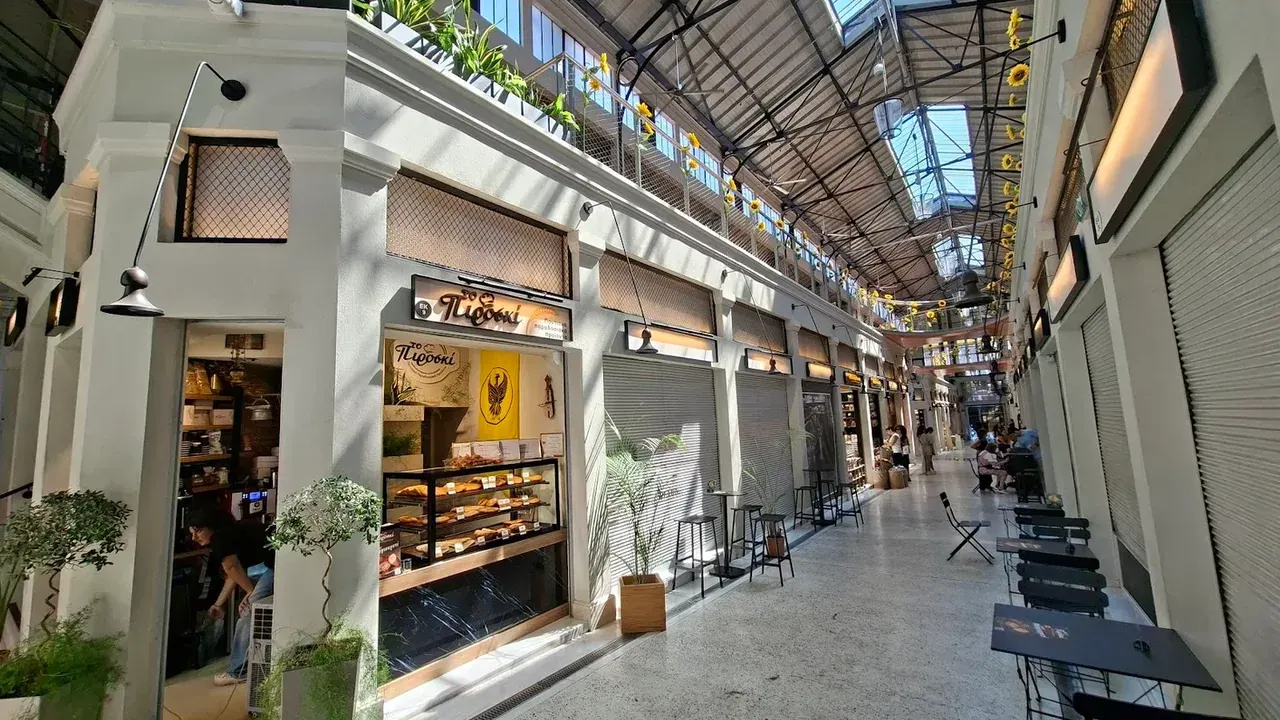 The glass-roofed arcade of the renovated Modiano Market in Thessaloniki, lined with small shopfronts, potted plants and café tables