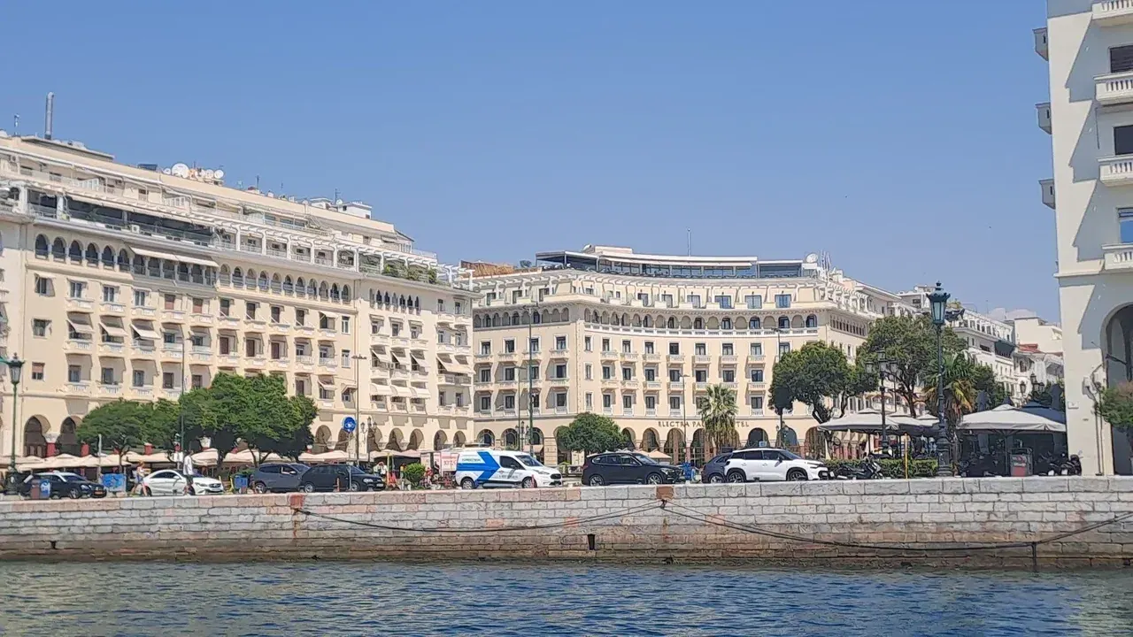 A waterfront hotel building with cafe terraces and parked cars, seen from across the water in central Thessaloniki