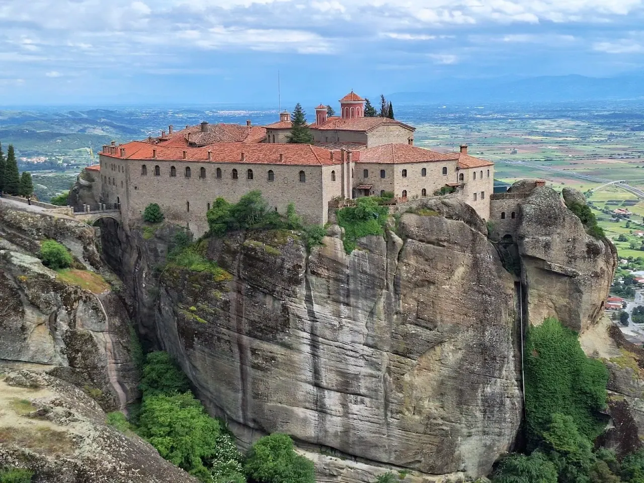 A large Meteora monastery complex spread across a broad sandstone rock above the green valley