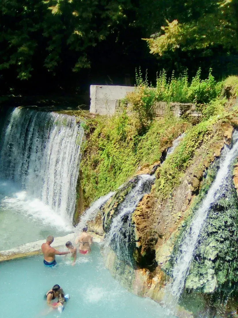 Bathers in a steaming turquoise thermal pool beneath a waterfall at Pozar