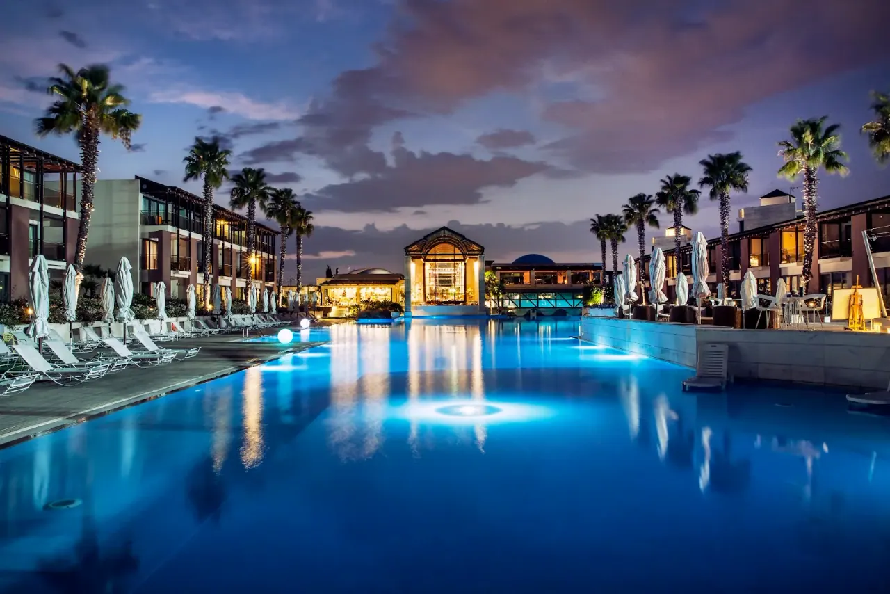 The illuminated resort pool at Hotel Nikopolis Thessaloniki at dusk, flanked by palm trees