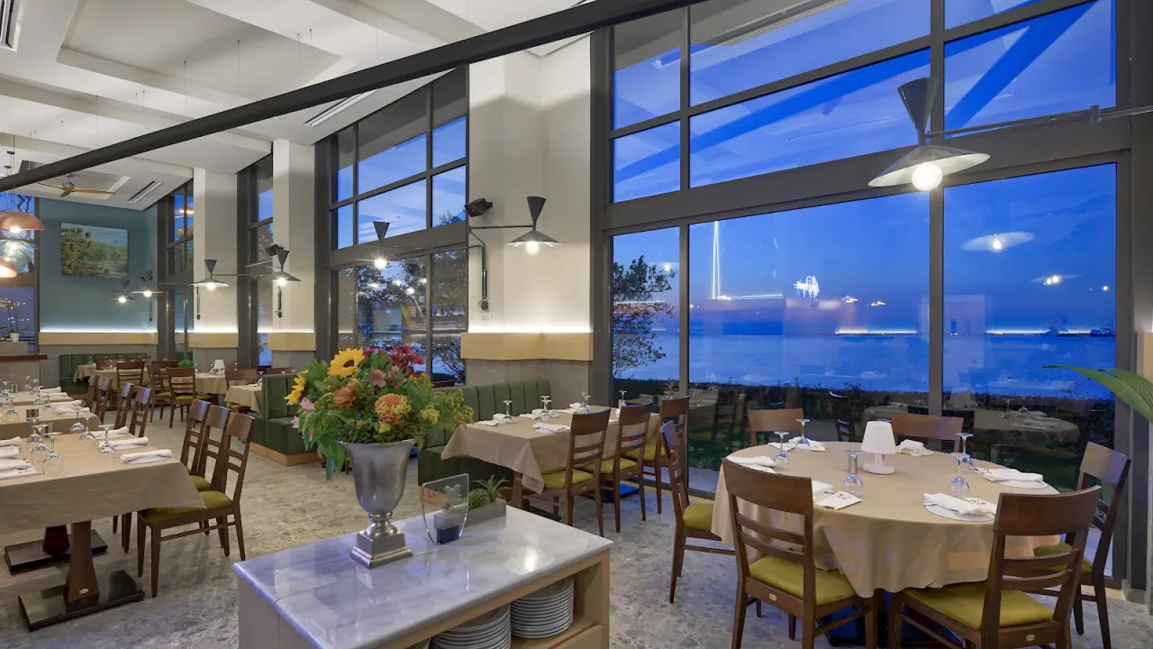 The high-ceilinged dining room of Nea Diagonios at dusk, tall windows looking over the Thermaic Gulf, Kalamaria, Thessaloniki