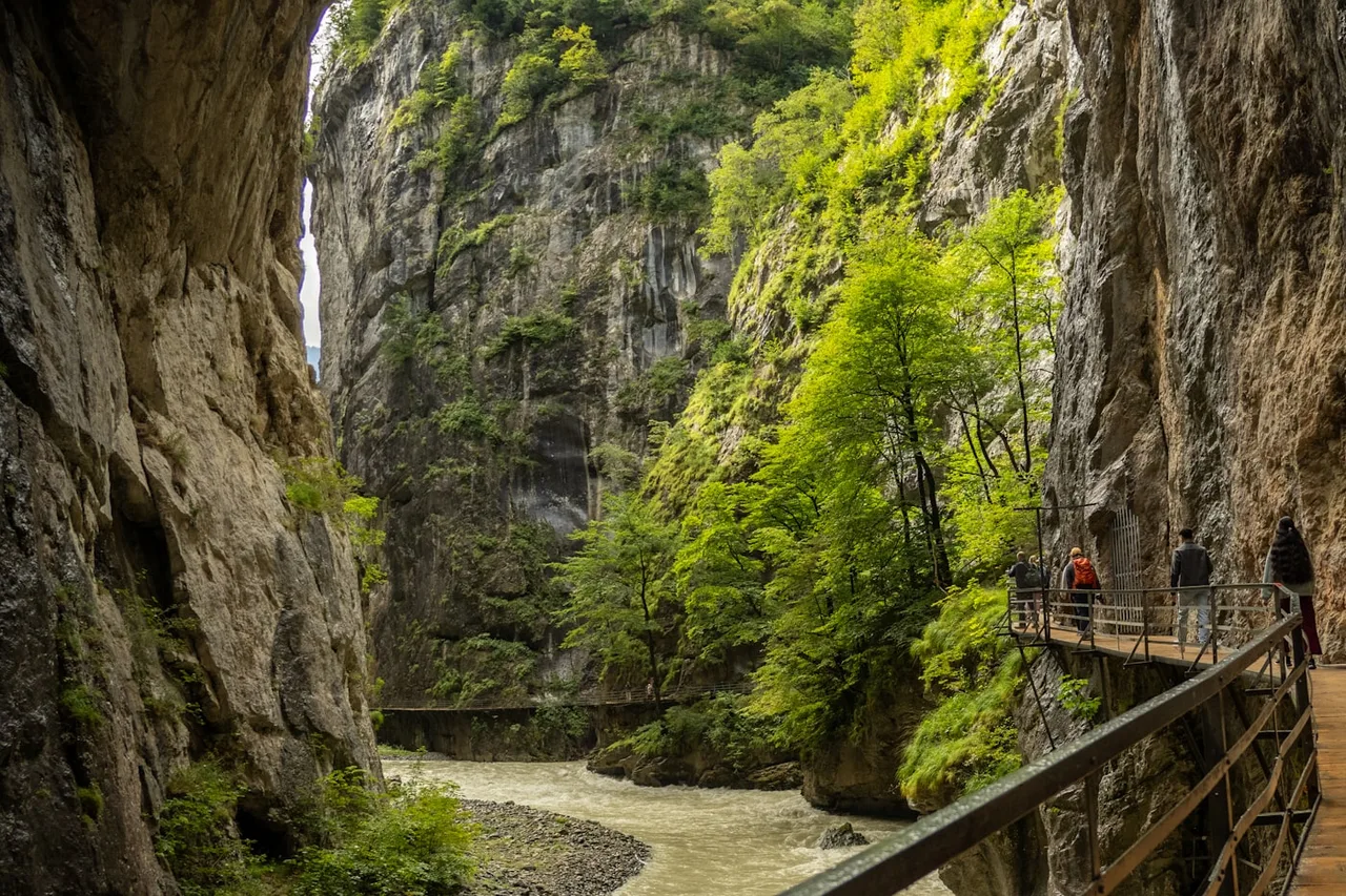 A railed walkway following a river through a steep gorge beneath limestone cliffs, like the Enipeas Gorge on Mount Olympus
