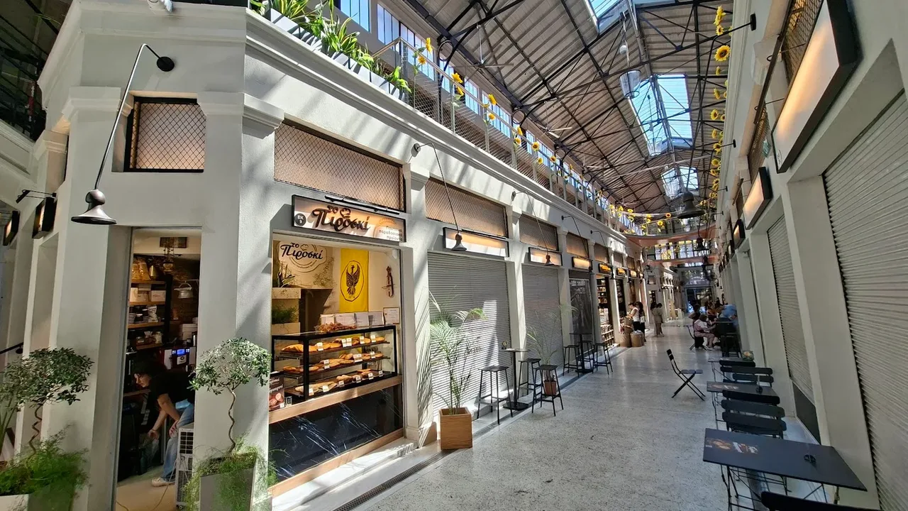 The glass-roofed arcade of the renovated Modiano Market in Thessaloniki, lined with small shopfronts, potted plants and café tables