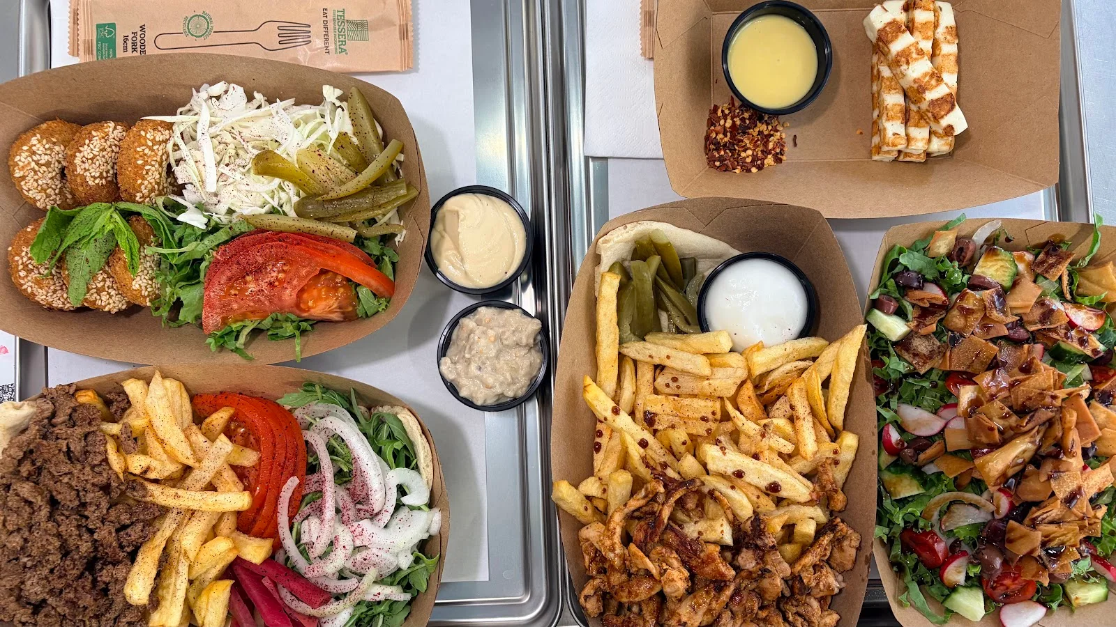 An overhead spread of Middle Eastern trays at Laziza — sesame falafel, chicken and fries, fattoush salad, grilled halloumi and dips, Thessaloniki